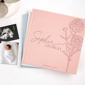 May include: A pink baby memory book with the name "Sophie Georgia" in script, a floral design, and the date "November 14, 2025." The book is stacked with other books and photos of a baby and an ultrasound.