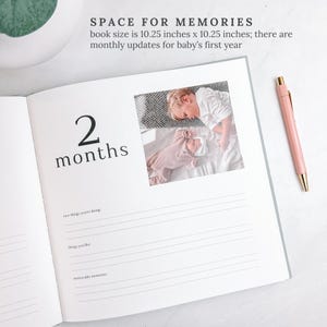 May include: Open baby memory book with a photo of a baby and a child. The book is open to a page that says "2 months". A pink pen with gold accents rests on the page. The book size is 10.25 inches x 10.25 inches.