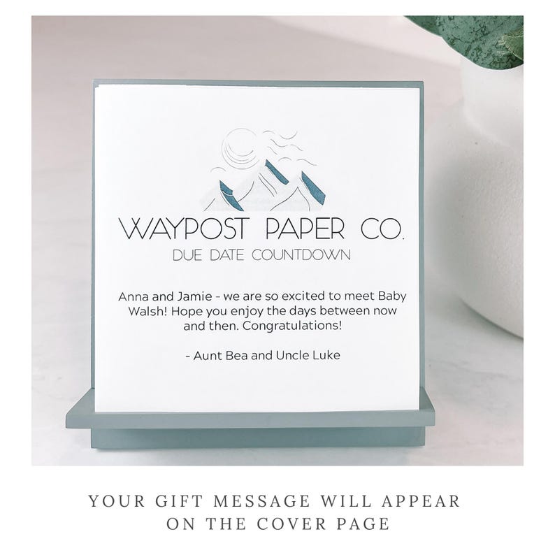May include: A Waypost Paper Co. due date countdown card. The card has a mountain logo and text that reads "Anna and Jamie - we are so excited to meet Baby Walsh!" The card is displayed on a light blue stand.