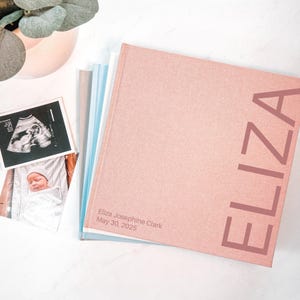May include: A stack of baby memory books with a pink cover. The word "ELIZA" is printed vertically on the cover. The book is next to a sonogram and a photo of a newborn baby. The text "Eliza Josephine Clark May 30, 2025" is printed on the cover.