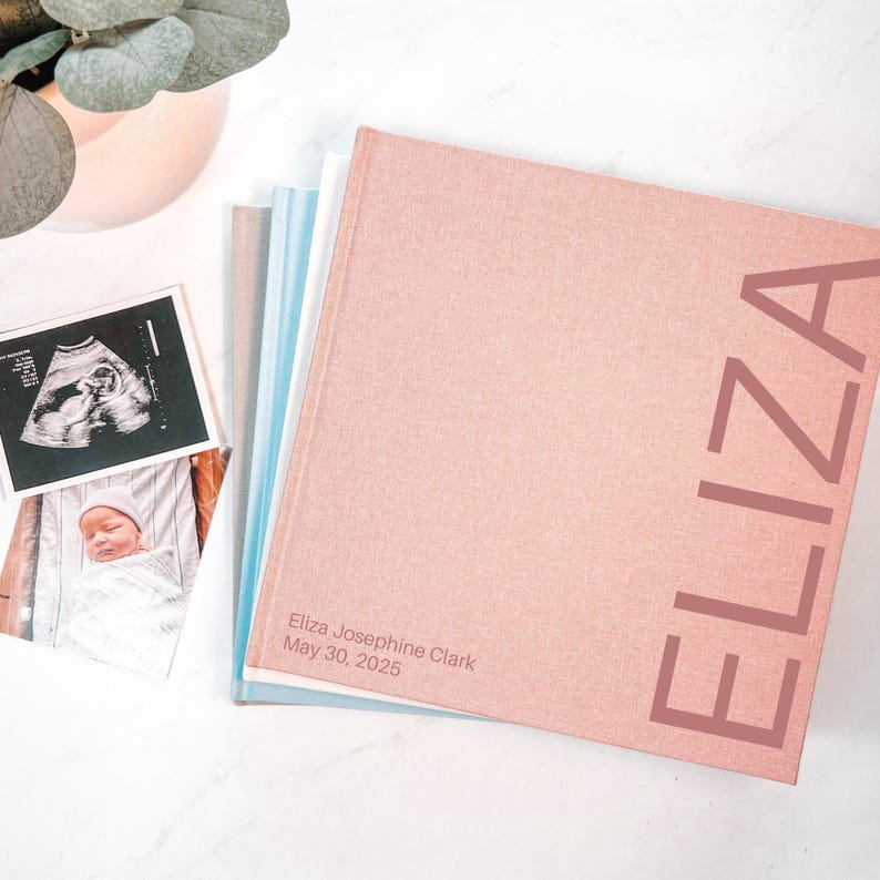 May include: A stack of baby memory books with a pink cover. The word "ELIZA" is printed vertically on the cover. The book is next to a sonogram and a photo of a newborn baby. The text "Eliza Josephine Clark May 30, 2025" is printed on the cover.