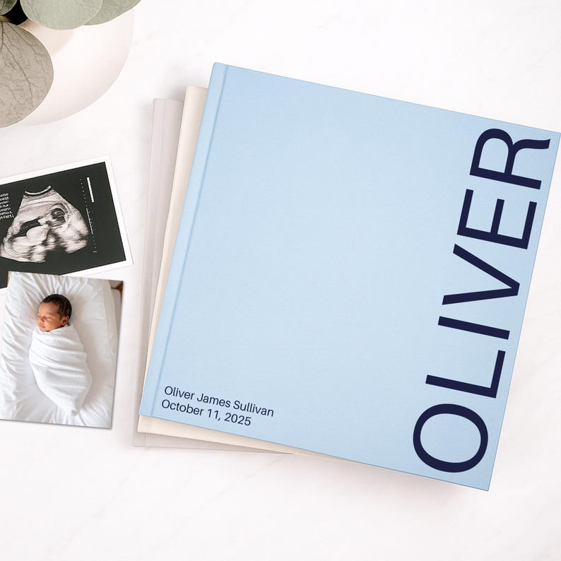 May include: A light blue baby memory book with the name "OLIVER" printed vertically in navy blue. The book also includes the name "Oliver James Sullivan" and the date "October 11, 2025". Beside the book are baby ultrasound and newborn photos.