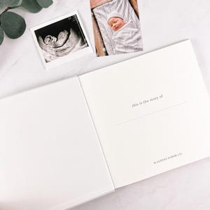 May include: An open, white baby memory book with the text "this is the story of" and "WAYPOST PAPER CO." printed on the right page. A sonogram and a photo of a newborn baby are on the top.