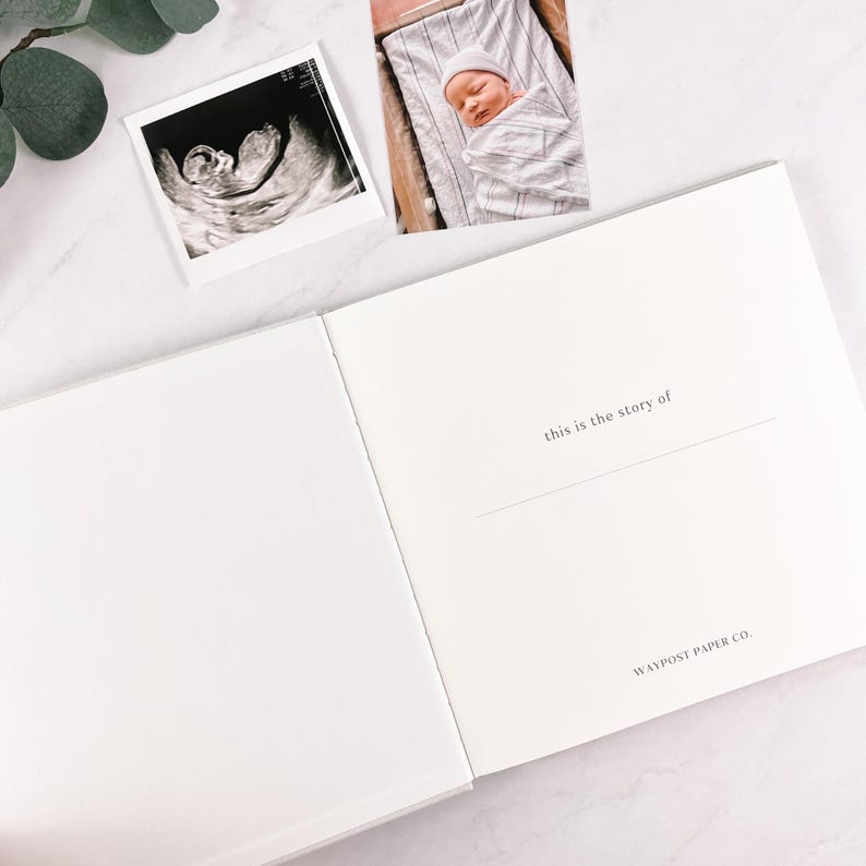 May include: An open, white baby memory book with the text "this is the story of" and "WAYPOST PAPER CO." printed on the right page. A sonogram and a photo of a newborn baby are on the top.