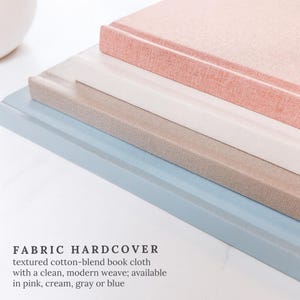 May include: A stack of fabric hardcover books in pink, cream, grey, and blue. The books have a textured cotton-blend book cloth with a clean, modern weave. The text "FABRIC HARDCOVER" is visible.