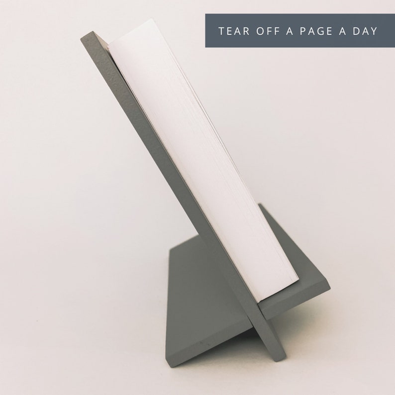 May include: A gray wooden desk calendar holder with a stack of white pages. The holder is angled, with the text "TEAR OFF A PAGE A DAY" in a dark gray rectangle at the top.
