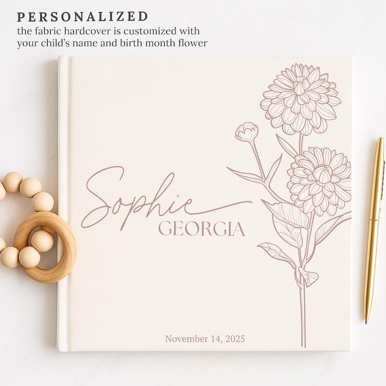 May include: A personalised fabric hardcover book featuring a floral design and the name "Sophie" and "Georgia" in a script font. The date "November 14, 2025" is printed at the bottom. A wooden teething ring and gold pen are next to the book.