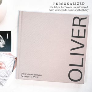 May include: A personalized fabric hardcover book with the name "OLIVER" printed vertically in large black letters. The book's cover is a light beige color. The text "Oliver James Sullivan October 11, 2025" is printed on the bottom of the book.