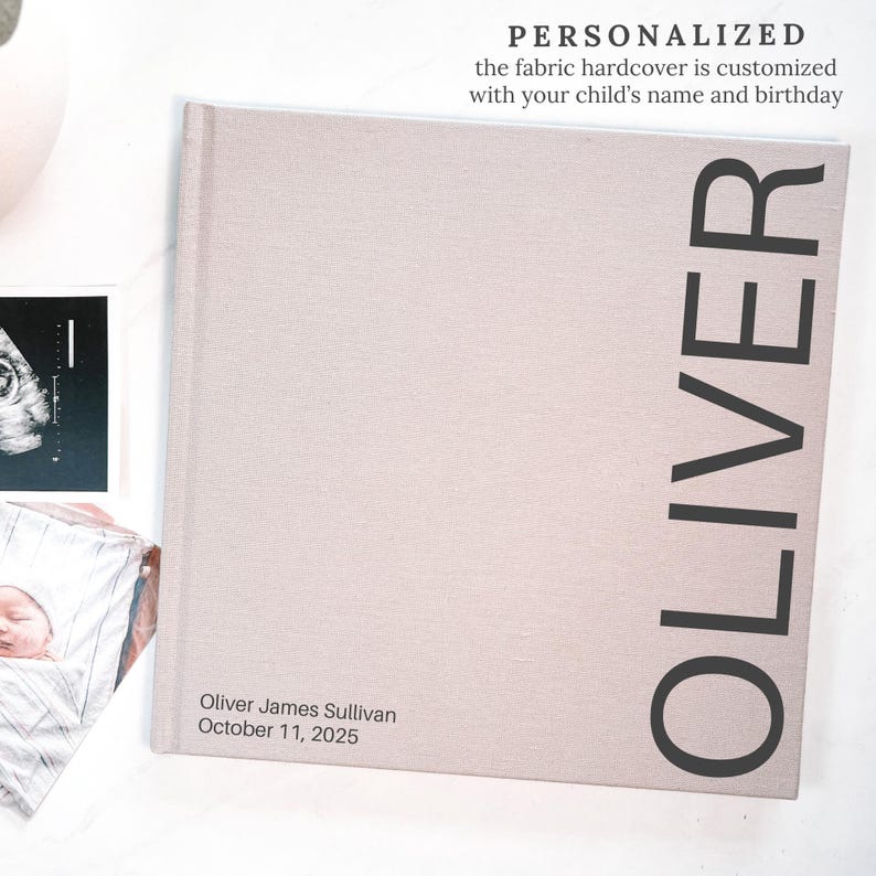 May include: A personalized fabric hardcover book with the name "OLIVER" printed vertically in large black letters. The book's cover is a light beige color. The text "Oliver James Sullivan October 11, 2025" is printed on the bottom of the book.