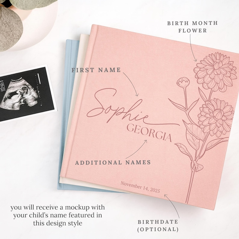 May include: A pink baby memory book with the name "Sophie" and "Georgia" printed on the cover, featuring a floral design. The book includes the optional birthdate of November 14, 2025. An ultrasound image is visible.
