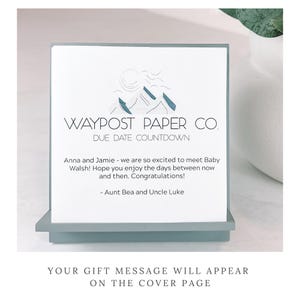 May include: A Waypost Paper Co. due date countdown card. The card has a mountain logo and text that reads "Anna and Jamie - we are so excited to meet Baby Walsh!" The card is displayed on a light blue stand.
