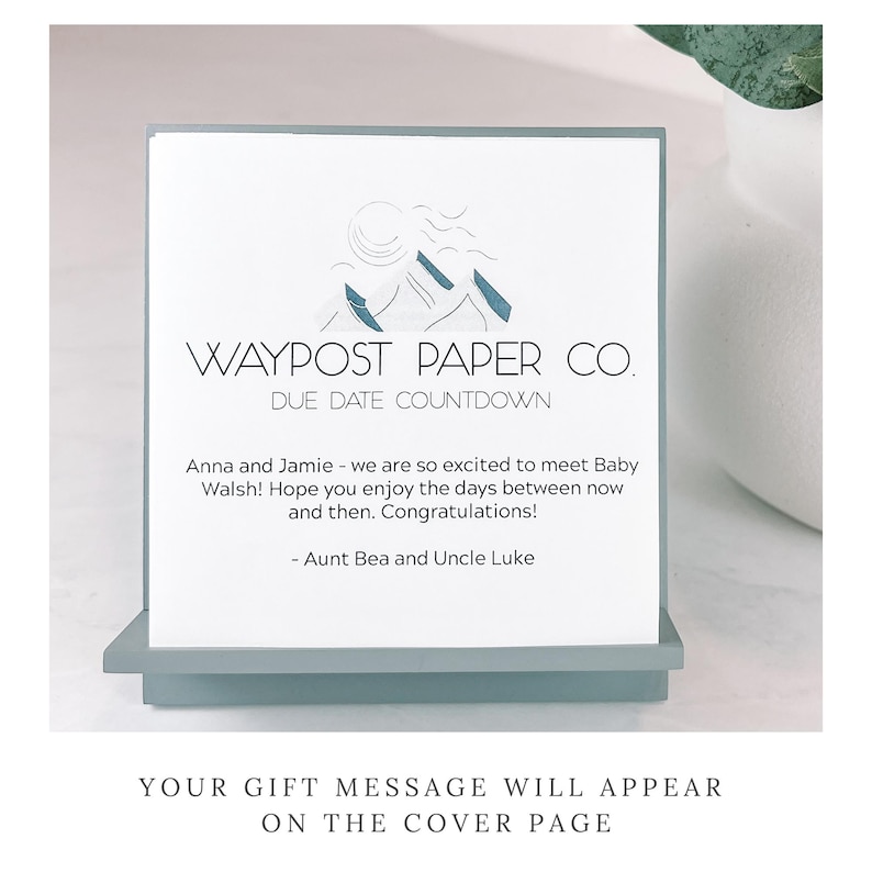 May include: A Waypost Paper Co. due date countdown card. The card has a mountain logo and text that reads "Anna and Jamie - we are so excited to meet Baby Walsh!" The card is displayed on a light blue stand.