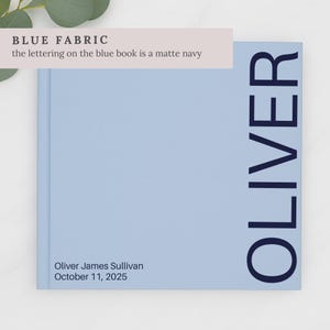 May include: A light blue book with the name "OLIVER" printed vertically in navy blue. The book also has the name "Oliver James Sullivan" and the date "October 11, 2025" printed on the bottom left corner. The text "BLUE FABRIC" is printed at the top.