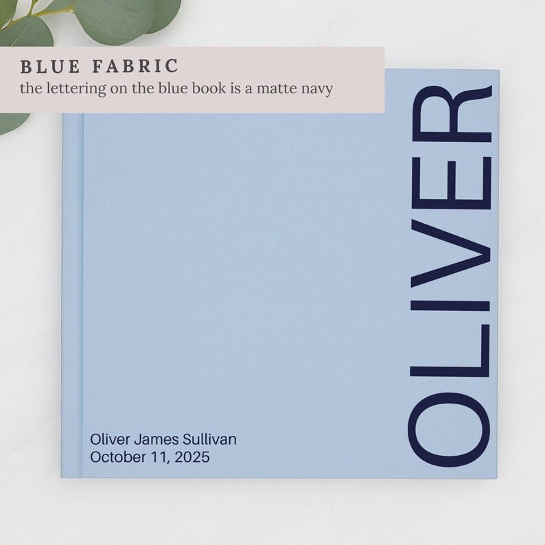 May include: A light blue book with the name "OLIVER" printed vertically in navy blue. The book also has the name "Oliver James Sullivan" and the date "October 11, 2025" printed on the bottom left corner. The text "BLUE FABRIC" is printed at the top.