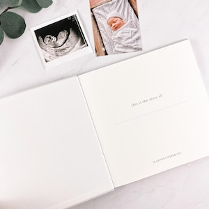 May include: A white baby memory book, open to a page with the text "this is the story of" and "WAYPOST PAPER CO.". A sonogram and a photo of a newborn baby are placed above the book.