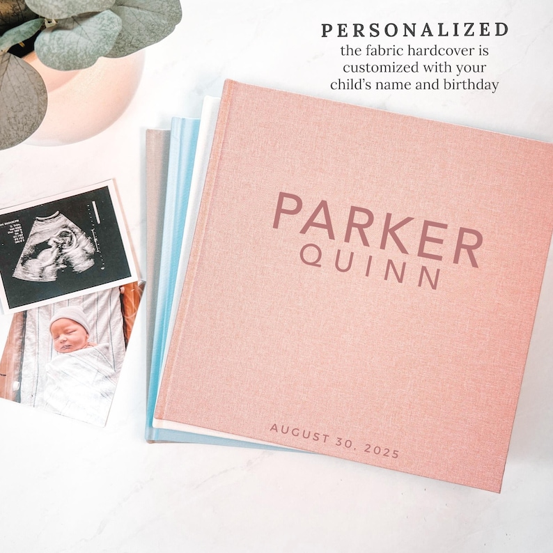 May include: A personalized fabric hardcover book in a soft pink hue, featuring the name "PARKER QUINN" and the date "AUGUST 30, 2025." The book is stacked with other books and next to baby photos and a sonogram.