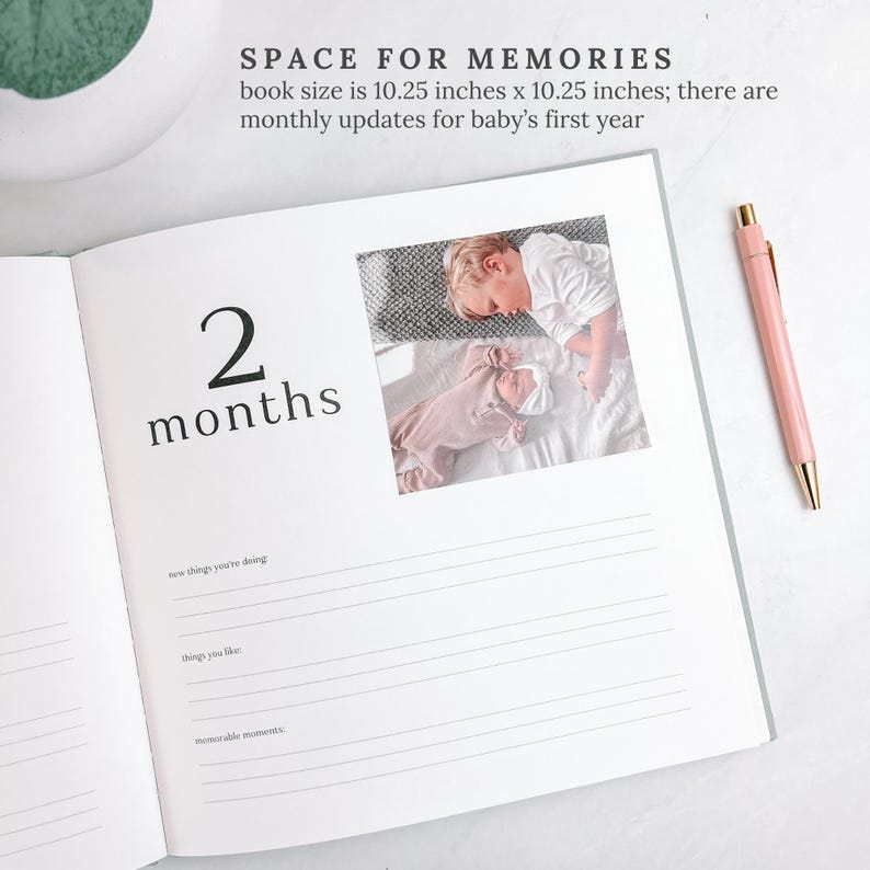 May include: Baby memory book open to a page with the text "2 months". A photo of a baby and a child is in the upper right corner. A pink pen with gold detailing lies on the page. The book is 26 cm x 26 cm.