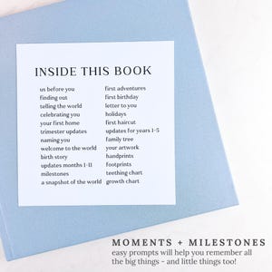 May include: A light blue baby memory book with a white label that reads "INSIDE THIS BOOK" with prompts like "first birthday" and "milestones." The bottom of the book reads "MOMENTS + MILESTONES".