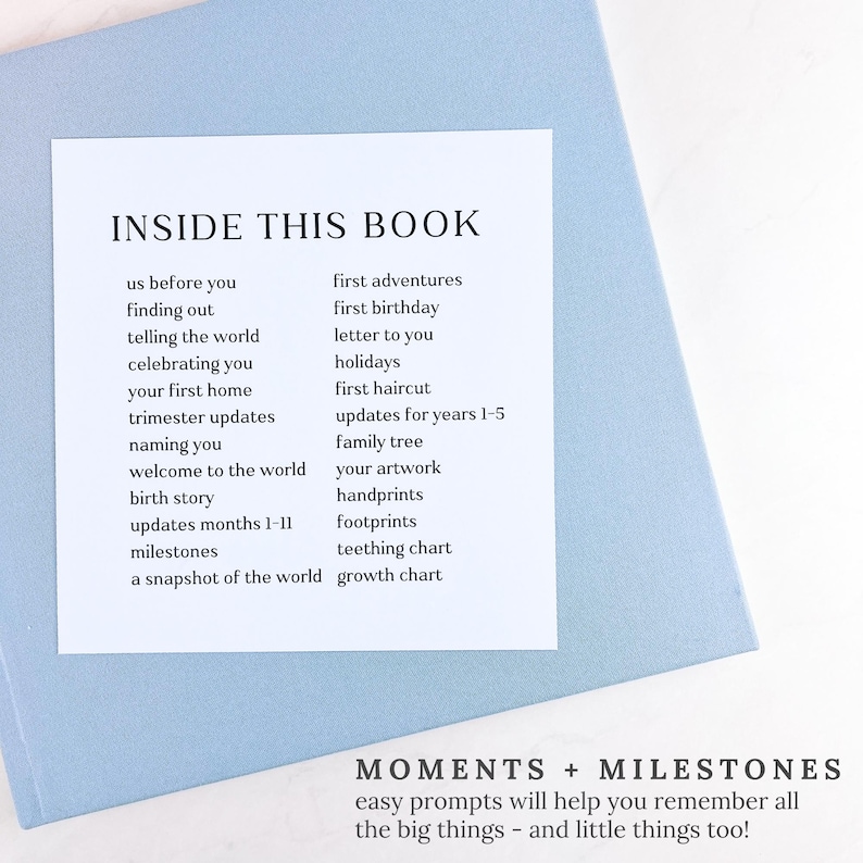 May include: A light blue baby memory book with a white label that reads "INSIDE THIS BOOK" with prompts like "first birthday" and "milestones." The bottom of the book reads "MOMENTS + MILESTONES".