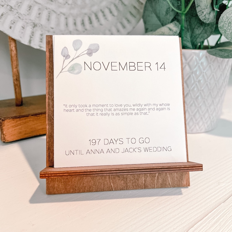 K&ouml;nnte beinhalten: Ein Holz-Schreibtischkalender zeigt "NOVEMBER 14" mit einem floralen Design. Der Kalender enth&auml;lt ein Zitat und den Text "197 DAYS TO GO UNTIL ANNA AND JACK'S WEDDING". Der Kalender ist aus hellbraunem Holz.
