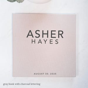 May include: A light gray book with the name "ASHER HAYES" in charcoal lettering. The date "AUGUST 30, 2025" is printed below the name. The book has a textured cover and is on a white surface.