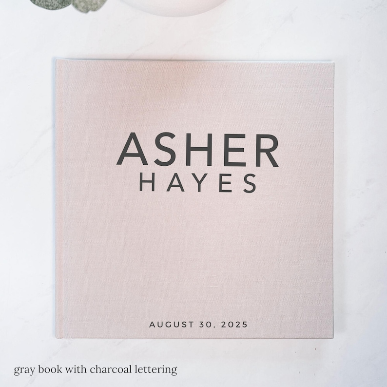 May include: A light gray book with the name "ASHER HAYES" in charcoal lettering. The date "AUGUST 30, 2025" is printed below the name. The book has a textured cover and is on a white surface.