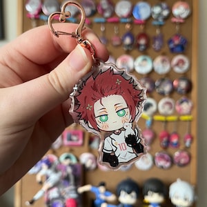 May include: A clear acrylic keychain with an anime-style character with red hair and green eyes, holding a white cat. The character wears a white jersey with the number 10. The keychain has a rose gold heart-shaped clasp.