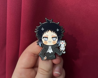 May include: Enamel pin depicting a cartoon character with black hair, blue highlights, and a black coat. The character is holding a white cat. The pin has a black and white colour scheme with blue and peach accents. The pin is held in a hand against a burgundy background.