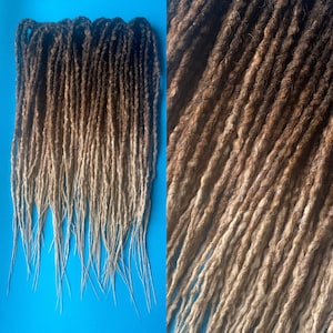 May include: A set of ombre brown and blonde synthetic dreadlocks. The dreadlocks are long and thick, with a dark brown color at the top that fades to a light blonde color at the bottom.