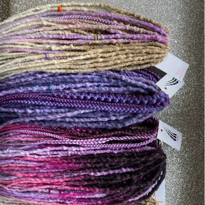 May include: Three bundles of synthetic dreadlocks in shades of purple, pink, and beige. The dreadlocks are arranged in a stack, with the beige dreadlocks on top, followed by the purple dreadlocks, and then the pink dreadlocks. The dreadlocks are all about 2 feet long.