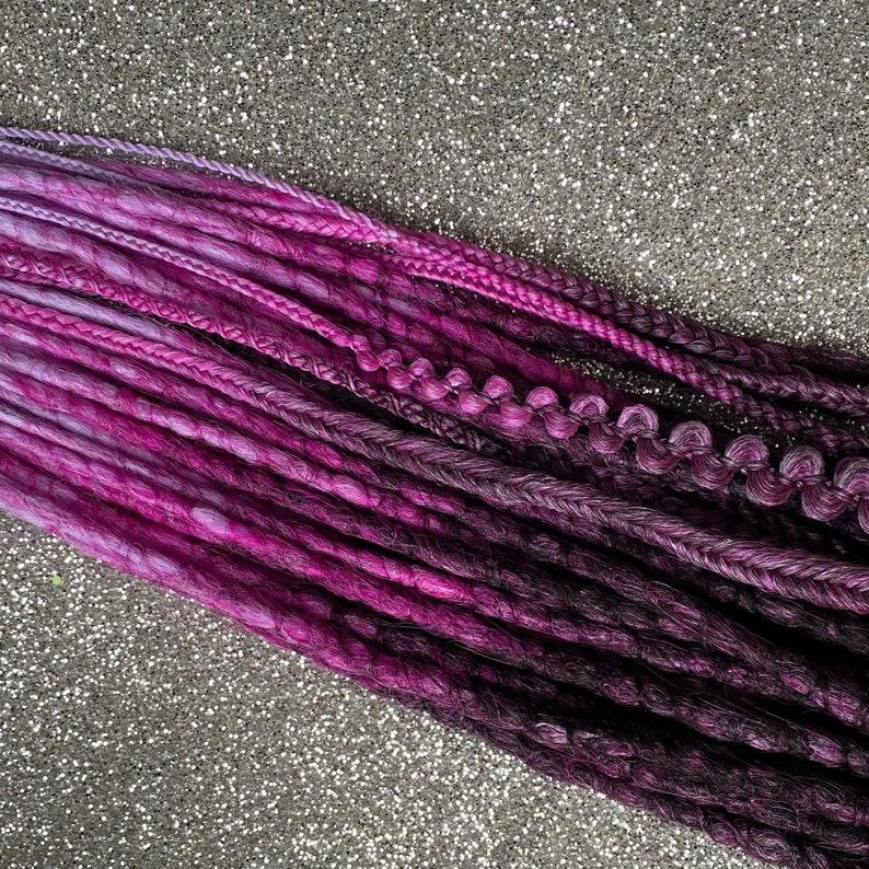May include: A set of purple and black synthetic dreadlocks. The dreadlocks are ombre, with the purple fading to black. The dreadlocks are arranged in a row, with the purple ends at the top and the black ends at the bottom.