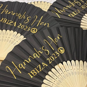 May include: A collection of decorative folding fans. Some fans are black, others white, with gold text that reads "Bride to Be IBIZA 2023" and "Hannah's Hen IBIZA 2023". Each fan has a peace symbol.
