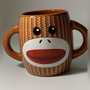 May include: A brown ceramic coffee mug with a monkey face design. The monkey has a white face with a red mouth and brown knitted eyebrows.