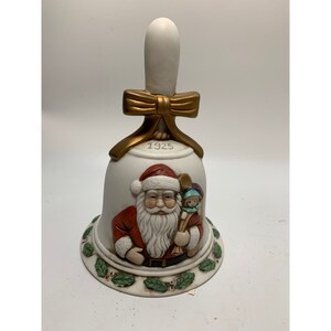 May include: White porcelain Christmas bell with a gold bow and handle. The bell features a raised image of Santa Claus holding a doll and the year 1925.