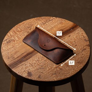 May include: A brown leather glasses case, measuring 7.1 inches long and 3.1 inches wide, with a button closure. The case has a decorative emblem and is displayed on a wooden stool.