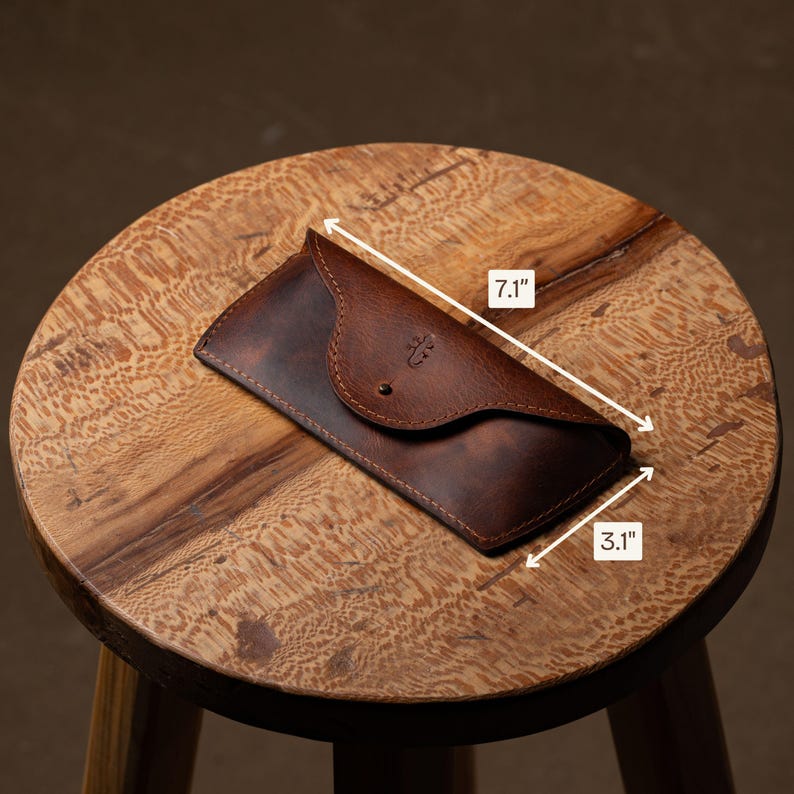 May include: A brown leather glasses case, measuring 7.1 inches long and 3.1 inches wide, with a button closure. The case has a decorative emblem and is displayed on a wooden stool.