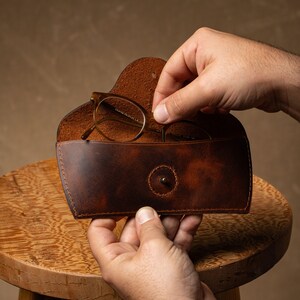 May include: A brown leather glasses case with a snap closure. The case is open, revealing a pair of eyeglasses. The case has a distressed finish and is being held by a person.