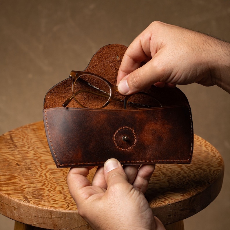 May include: A brown leather glasses case with a snap closure. The case is open, revealing a pair of eyeglasses. The case has a distressed finish and is being held by a person.
