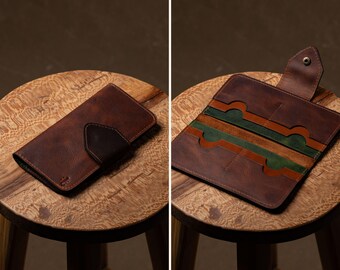 Puede incluir: Cartera de cuero marrón, cerrada y abierta, expuesta sobre un taburete de madera. La cartera cerrada tiene una solapa marrón oscuro. La cartera abierta revela ranuras para tarjetas con detalles en verde y naranja. La cartera es de cuero texturizado.