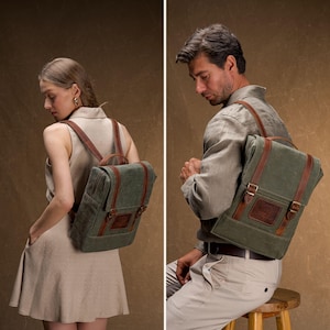 May include: A green canvas backpack with brown leather straps and accents. The backpack has a rectangular shape with a flap closure and a small front pocket. The bag is being worn by a person.
