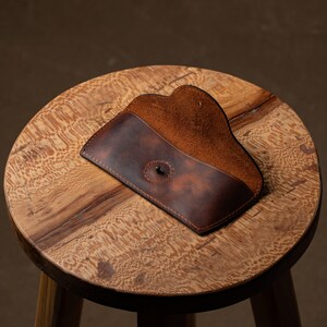 May include: A brown leather glasses case with a snap closure, open to reveal a lighter brown interior. The case is resting on a small, round wooden stool with a natural wood grain finish. The background is a muted brown.