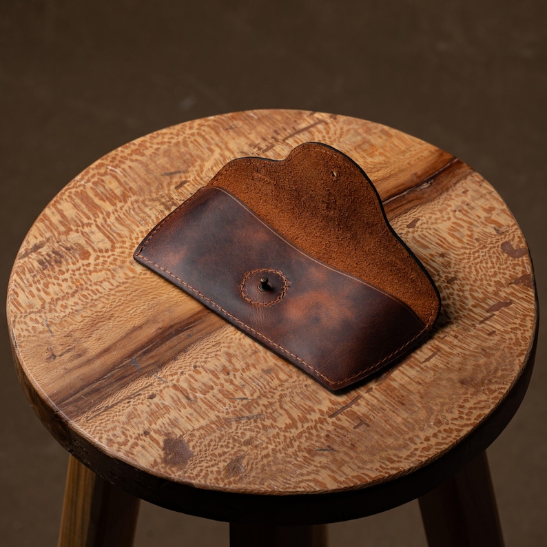 May include: A brown leather glasses case with a snap closure, open to reveal a lighter brown interior. The case is resting on a small, round wooden stool with a natural wood grain finish. The background is a muted brown.