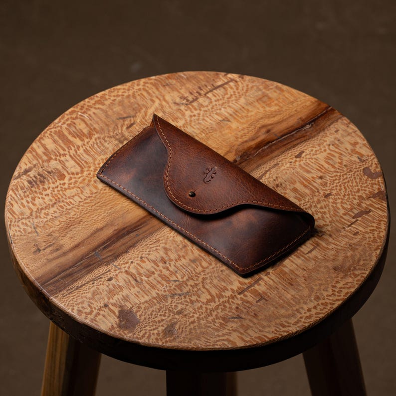 May include: A brown leather glasses case with a curved flap and a small button closure. The case has a textured surface and a decorative emblem. It rests on a wooden stool with a natural wood grain finish.