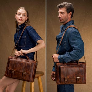 May include: Two people are shown with brown leather briefcases. The briefcases have a rectangular shape with a top handle and a shoulder strap. The briefcases have two buckles on the front. The leather has a distressed look.
