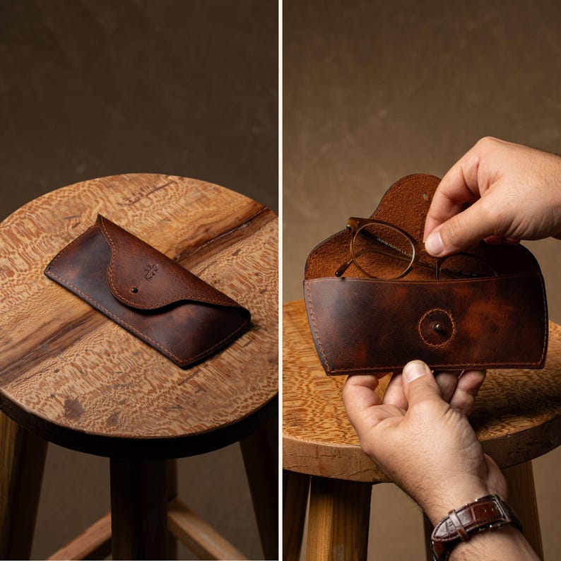 May include: A brown leather glasses case with a snap closure, shown open and closed. The case is on a wooden stool. The glasses case is designed to protect eyeglasses. The case has a logo embossed on the front.