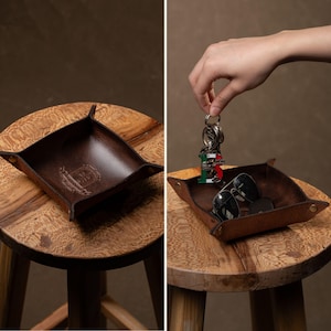 May include: A brown leather catchall tray with a square shape and raised edges, resting on a wooden stool. The tray has a stamped design in the center. The second image shows the tray holding sunglasses and a set of keys.