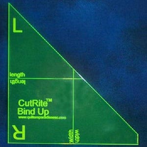 以下が含まれることがあります： 緑色のプラスチック製の三角定規で、「CutRite Bind Up」と「www.quiltinpatchladies.com」の文字が書かれています。定規には長さの目盛りと幅の目盛りがあります。
