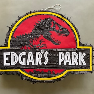 May include: A black and yellow Jurassic Park-themed piñata with a red background and a black dinosaur silhouette. The text "EDGAR'S PARK" is printed in white on the piñata.