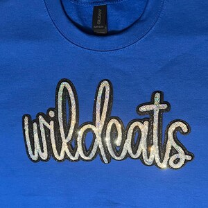 May include: A royal blue sweatshirt featuring the word "wildcats" in a sequined, cursive font. The text is outlined in black and has a shimmering, iridescent effect. The sweatshirt is a solid colour.