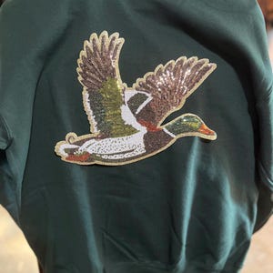 Può includere: Felpa verde scuro con un'anatra volante di paillettes. L'anatra presenta dettagli marroni, dorati, verdi, bianchi e rossi. La felpa è di colore verde scuro.